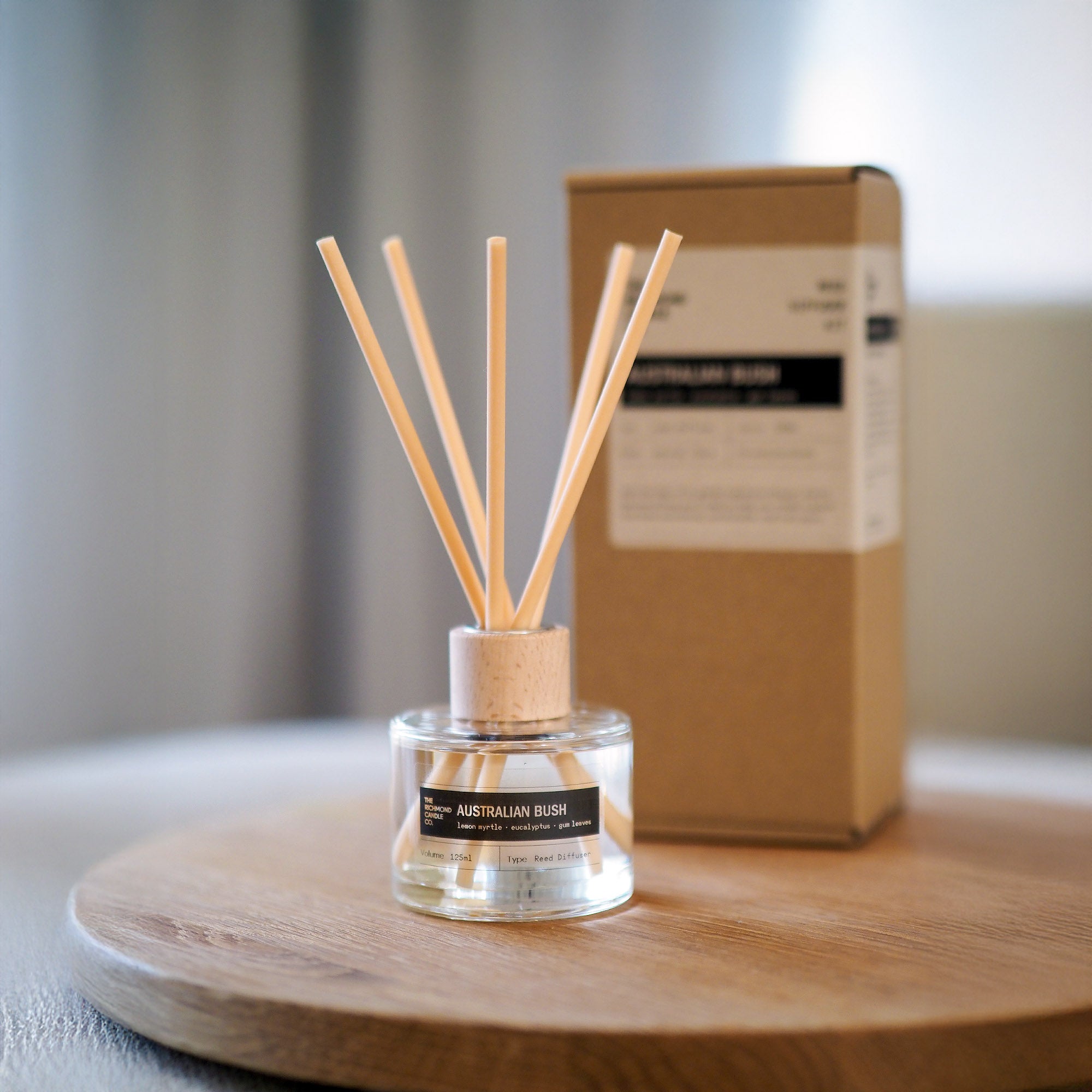 Reed diffuser with wooden sticks in a clear container on a wooden surface, with the diffuser box in the background